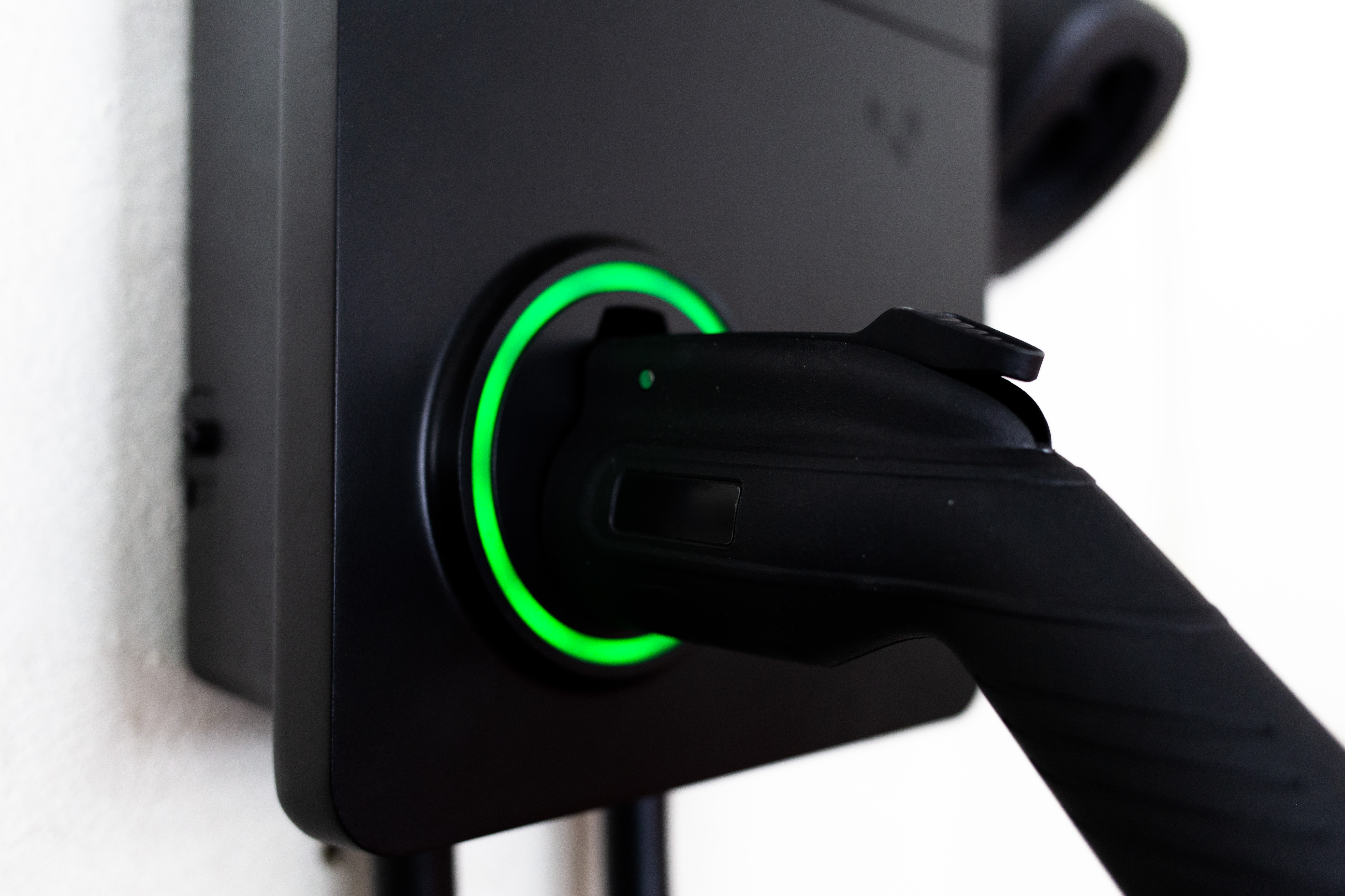 Close-up of a black electric vehicle wall charger with a charging plug inserted, surrounded by a glowing green LED ring indicating active charging