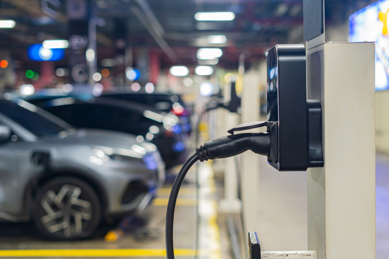 An electric vehicle charging cable plugged into a wall-mounted charger in a parking garage, with a parked car blurred in the background under overhead lights.